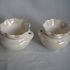 May include: Two pearlescent white ceramic bowls with a scalloped rim. Each bowl has a smaller, matching bowl nested on top. The larger bowls have small handles on either side. The bowls have an iridescent sheen.