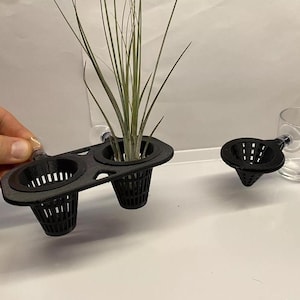 May include: Black plastic plant holders with suction cups, designed for hanging plants. Two holders are attached together, and a third holder is separate. A small green plant is in one of the holders.