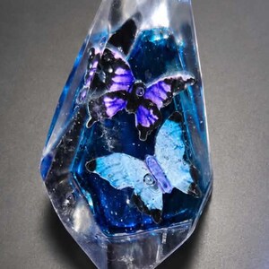 Könnte beinhalten: Ein klarer, facettierter, kristallförmiger Briefbeschwerer aus Kunstharz mit einem tiefblauen, glitzernden Inneren. Im Inneren sind zwei Schmetterlinge eingebettet: einer mit violetten und schwarzen Flügeln über einem hellblauen und schwarzen.
