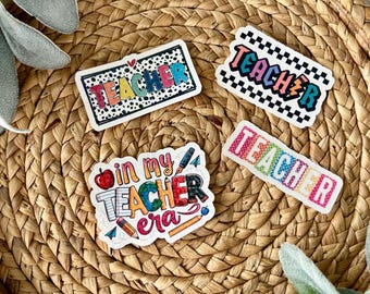 May include: Four colorful stickers with the word "TEACHER" and the phrase "in my TEACHER era". The stickers have various designs, including polka dots, a checkered pattern, and a rainbow color scheme. The stickers are on a woven, circular tray.