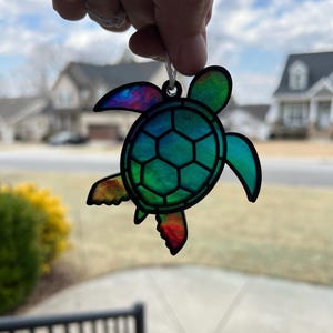 May include: A colorful sea turtle ornament with a black outline. The turtle's shell is a mosaic of green, blue, and purple hues. The flippers have red, orange, and yellow accents. A white string is attached for hanging.