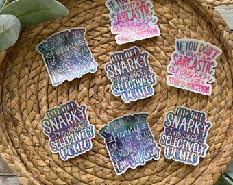 May include: A collection of colorful stickers with humorous phrases. The stickers feature text like "I'm not snarky, I'm just selectively polite" and "If you don't want a sarcastic answer, don't ask a stupid question." The stickers are arranged on a woven basket.