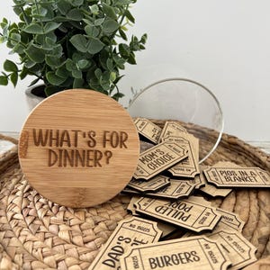 May include: A wooden lid with the text "WHAT'S FOR DINNER?" sits next to a glass jar filled with wooden tickets. The tickets have various dinner options like "BURGERS" and "PIGS IN A BLANKET". A small potted plant is in the background.