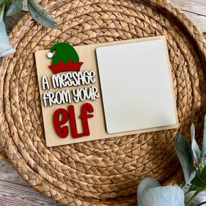 Puede incluir: Un letrero de madera con el texto "A message from your Elf" y un espacio en blanco para escribir. El letrero presenta un gorro de elfo verde con un borde rojo. El letrero está colocado sobre una cesta tejida.