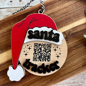May include: Wooden Christmas ornament featuring a red Santa hat design with the words "santa tracker" in black. The ornament includes a QR code and star accents, with a black and white string for hanging.