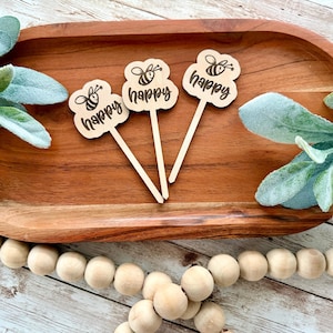 May include: Three wooden bee-themed toppers with the word "happy" and a bee illustration. The toppers are on a wooden tray with greenery and a wooden bead garland. The toppers are light brown with black text.