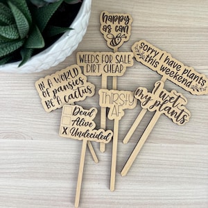 May include: Set of six wooden garden stakes with humorous sayings, including "Happy as can", "Weeds for sale dirt cheap", "In a world of pansies be a cactus", "Sorry I have plants this weekend", "I wet my plants", and "Thirsty AF".