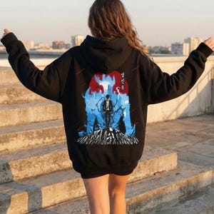 Puede incluir: Sudadera con capucha negra con un diseño gráfico en la espalda que presenta una figura en un paisaje azul y rojo. El diseño incluye texto coreano. La persona está de pie en unos escalones de hormigón.