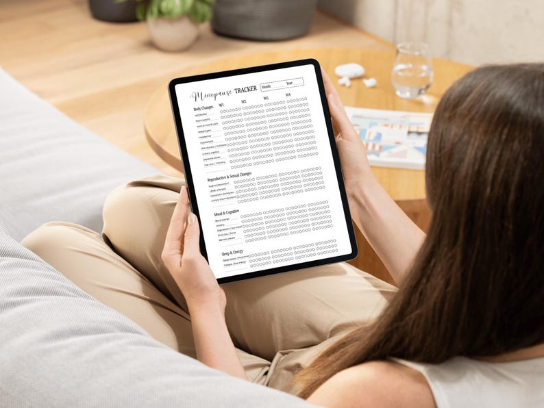 May include: A digital tablet displaying a "Menopause Tracker" with sections for body changes, reproductive changes, mood, and sleep. The tablet is held by a person, with the screen's text in a clear, readable font. The background includes a couch and a table.