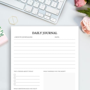 May include: A white Daily Journal with lined pages, the title "DAILY JOURNAL" in a dark gray font. The journal includes prompts for "1 MINUTE JOURNALING", "TOP 3 THINGS ABOUT TODAY", and "WHAT INSPIRED YOU THE MOST?" A keyboard, floral notebook, and flowers are in the background.