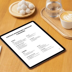 May include: A digital tablet displaying a "Capsule Wardrobe Checklist" for four seasons, with a stylus, a cup of coffee, a glass of water, and a plate of coconut macaroons on a wooden table. The checklist includes clothing categories.