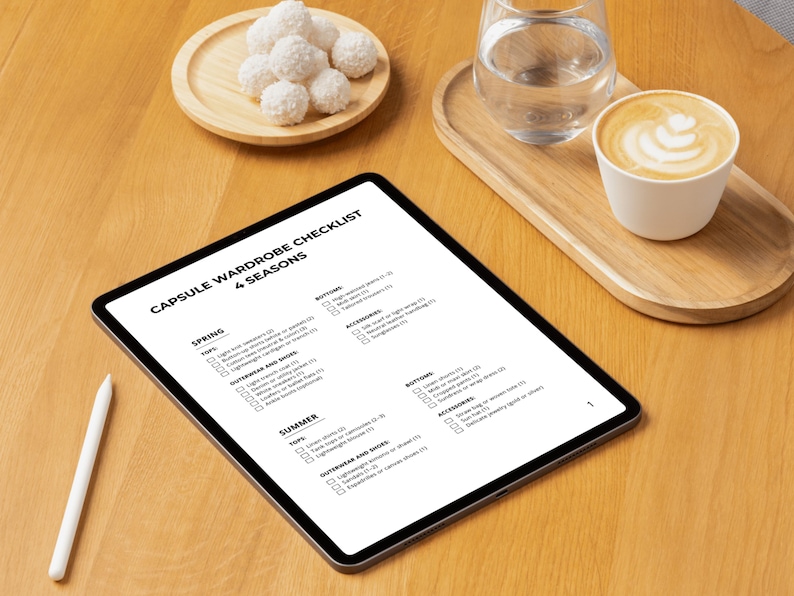 May include: A digital tablet displaying a "Capsule Wardrobe Checklist" for four seasons, with a stylus, a cup of coffee, a glass of water, and a plate of coconut macaroons on a wooden table. The checklist includes clothing categories.