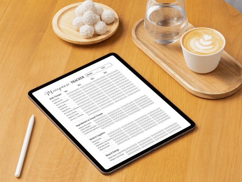 May include: A digital tablet displaying a "Menopause Tracker" form, alongside a white stylus. The image includes a wooden tray with a glass of water and a cup of coffee with latte art, and a small plate of coconut macaroons.