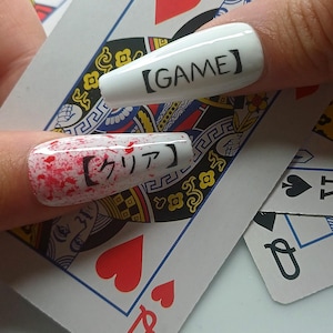 以下が含まれることがあります： 黒い文字で「GAME」と書かれた白い棺型のネイル。もう一方のネイルは、赤い飛沫模様と日本語のテキストが特徴です。ネイルは赤いハートの付いたトランプの上に表示されています。