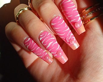 May include: Close-up of long, pink fingernails with a white zebra stripe design. Each nail has a small, pearl-like embellishment. The nails are adorned with gold rings.