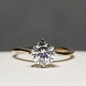 May include: A gold engagement ring featuring a large, round, clear gemstone. The ring's band has a curved design, and the gemstone is held in place by small prongs. The ring is shown on a white surface with a grey backdrop.