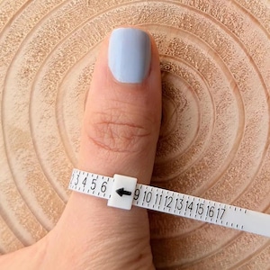 Op de afbeelding: Een witte ringmaat is om een duim met lichtblauwe nagellak gewikkeld. De maat heeft zwarte cijfers en een zwarte pijl die naar de maat wijst. De achtergrond is een lichtbruin houten oppervlak.