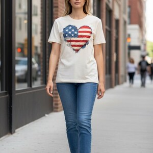 May include: A cream-colored t-shirt featuring a heart-shaped design of the American flag. The shirt is paired with blue jeans and white sneakers. The person is walking on a sidewalk in front of a brick building.