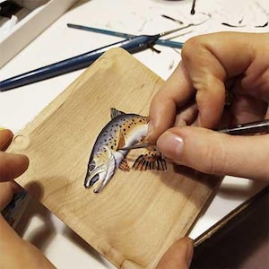 Può includere: Una mano dipinge dettagli su una piccola targa di legno con una rappresentazione realistica di una trota marrone.