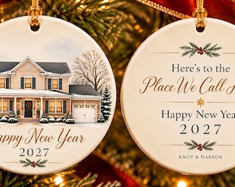 May include: A round white ceramic ornament featuring a two-story house illustration with snow, pine trees, and the text "Happy New Year 2027". The reverse side displays the text "Here's to the Place We Call Home Happy New Year 2027" with pine sprigs.