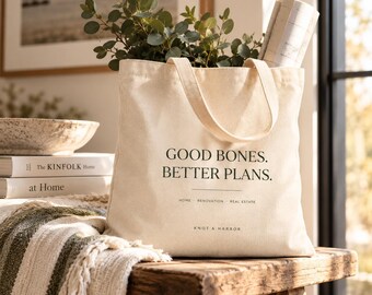 May include: A cream-colored canvas tote bag featuring the black printed text GOOD BONES. BETTER PLANS. HOME RENOVATION REAL ESTATE KNOT & HARBOR. The bag contains green leafy branches and a rolled paper document, with two sturdy fabric handles.
