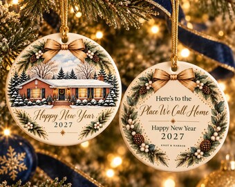 May include: Two round ceramic ornaments with gold hanging strings. The left ornament features a brick house in snow with the text Happy New Year 2027. The right ornament shows a pine wreath with the text Here's to the Place We Call Home.
