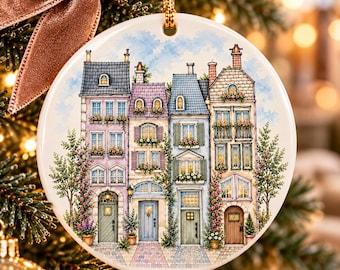 May include: A round white ceramic ornament featuring a detailed watercolor illustration of four multi-story townhouses with pastel facades, flower boxes, and climbing vines, suspended by a gold cord with a mauve velvet ribbon.