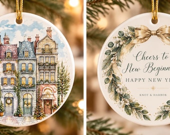 May include: A round white ceramic ornament featuring a detailed illustration of European-style townhouses on one side and a green wreath with the text Cheers to New Beginnings HAPPY NEW YEAR KNOT & HARBOR on the other side.