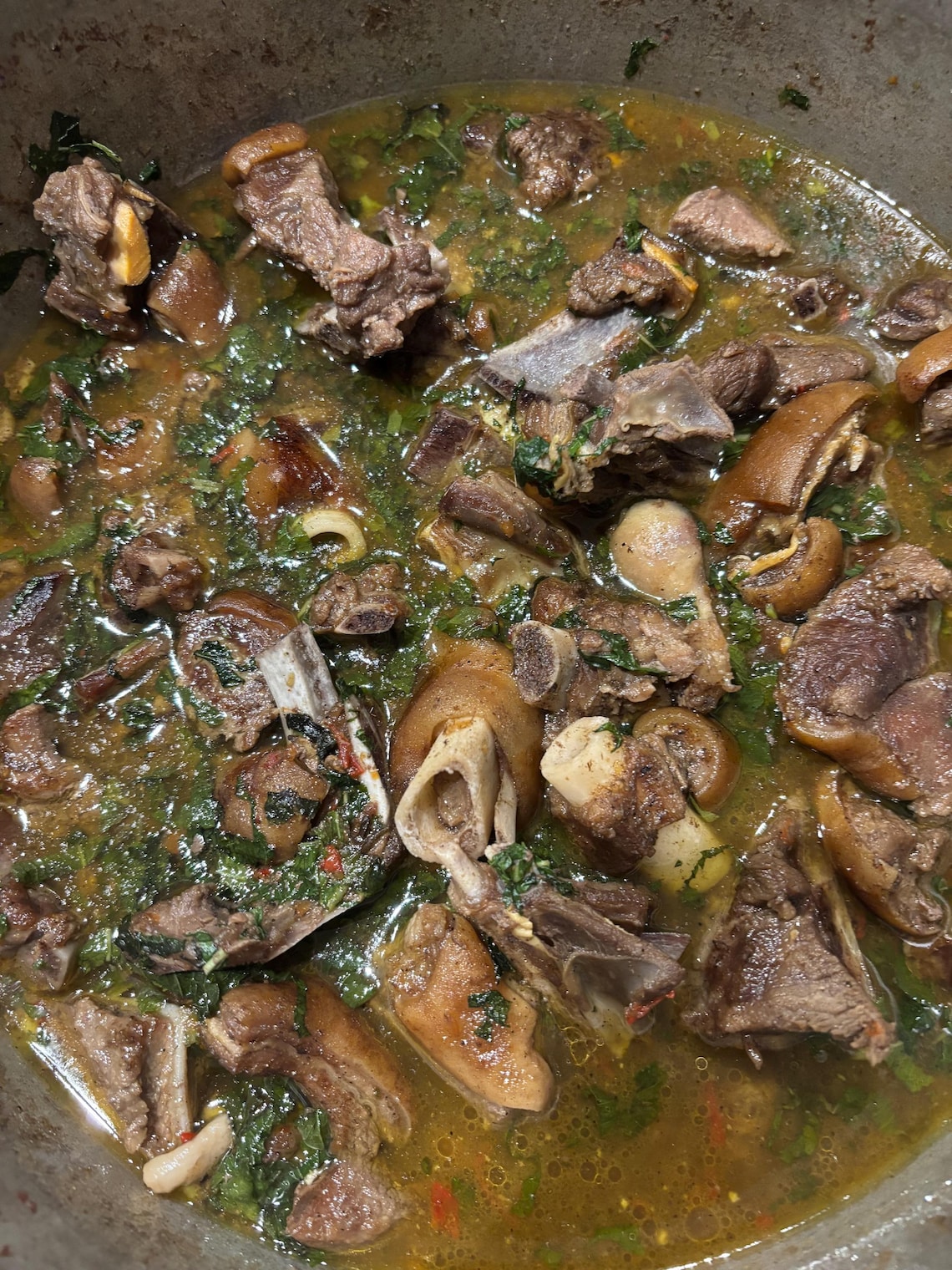 May include: A close-up of a pot of stew. The stew features various cuts of meat, including bone-in pieces and what looks like pig's trotters, in a broth with green leafy vegetables. The stew has a rich, appetising appearance.