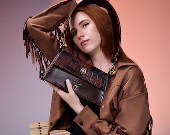 May include: A brown leather handbag with a crocodile-textured flap and a smooth leather body. The bag features a gold-coloured clasp and the brand name "SHLURA" is visible. The bag is held by a person wearing a brown jacket with fringe detailing.