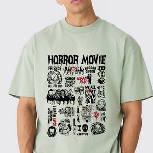 Peut inclure: T-shirt vert clair avec un motif noir. Le motif pr&eacute;sente les mots "HORROR MOVIE" et diverses r&eacute;f&eacute;rences de films d'horreur. Le motif comprend des images et du texte comme "FRIENDS", "HORROR QUEEN" et "WE ALL FLOAT DOWN HERE".
