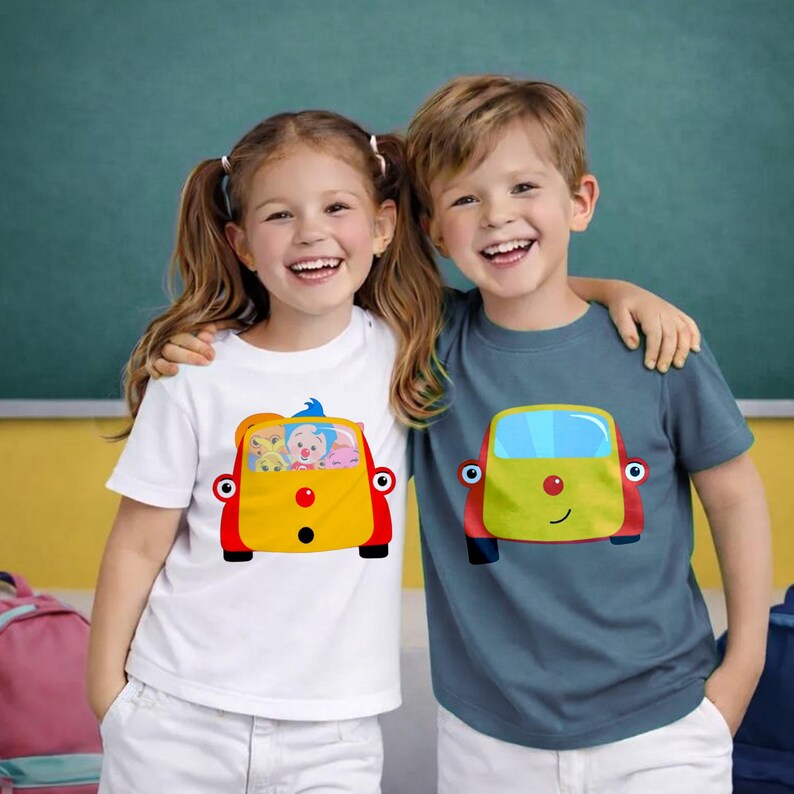 May include: Two children wearing t-shirts with cartoon bus designs. The t-shirts are white and blue-grey. The bus graphics are yellow, red, and blue, with smiling faces and cartoon characters inside. The children are smiling.