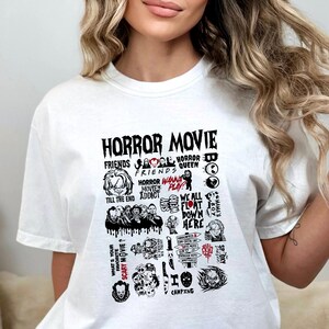 Peut inclure: T-shirt blanc avec un motif graphique noir repr&eacute;sentant les mots "HORROR MOVIE" et diverses images et phrases sur le th&egrave;me des films d'horreur. Le motif comprend des images de personnages et des citations de films d'horreur populaires.