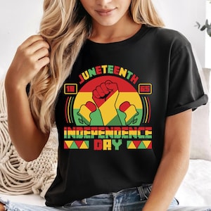 Puede incluir: Camiseta negra con un gráfico colorido del Día de la Independencia de Juneteenth. El diseño presenta un puño en alto en rojo, amarillo y verde, con el texto "Juneteenth Independence Day". La camiseta tiene cuello redondo clásico y mangas cortas.