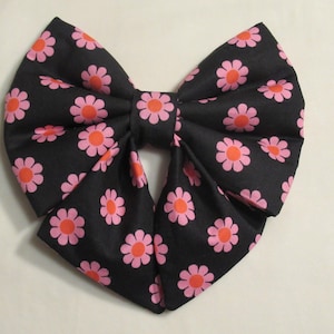 May include: A large, fabric bow tie with a black background and a repeating pattern of pink and orange flowers. The bow tie is tied in the center, creating a symmetrical shape. The flowers are evenly spaced across the fabric.