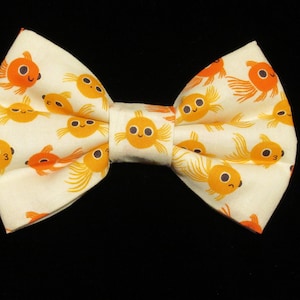 May include: A cream-colored bow tie featuring a repeating pattern of orange goldfish with black eyes. The bow tie is a classic shape, with a central knot and two symmetrical loops on either side. The background is black.