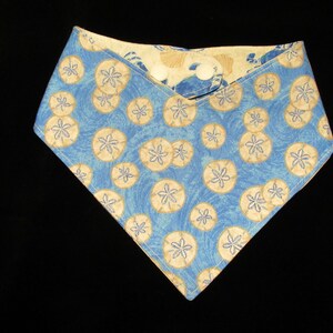 May include: A reversible pet bandana with a blue background and a pattern of beige sand dollars. The bandana is secured with a white button. The reverse side features a beige and white seashell pattern.