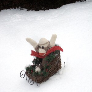 May include: A sock monkey wearing a red scarf sits in a wicker sled decorated with a green garland and red berries. The sled is on a snowy surface.
