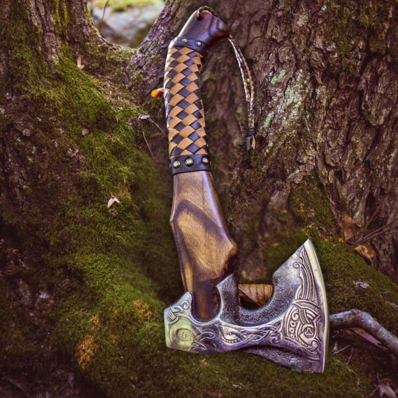 Puede incluir: Un hacha forjada a mano con un mango de madera envuelto en cuero marr&oacute;n. La cabeza del hacha tiene tallas intrincadas y una correa de cuero unida al mango.