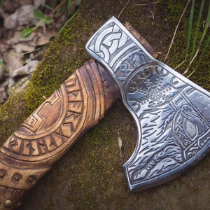 Puede incluir: Un hacha hecha a mano con un mango de madera tallado con intrincados patrones. La cabeza del hacha est&aacute; hecha de metal y presenta un dise&ntilde;o detallado de un &aacute;rbol y otros s&iacute;mbolos.