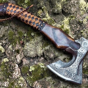 Puede incluir: Un hacha forjada a mano con un mango de madera envuelto en cuero marr&oacute;n. La cabeza del hacha presenta intrincados grabados de color plateado de un dise&ntilde;o de nudos vikingos.