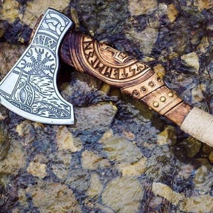 Puede incluir: Un hacha hecha a mano con mango de madera y cabeza de acero. La cabeza est&aacute; decorada con intrincados tallados, incluyendo un s&iacute;mbolo de una rueda solar y un p&aacute;jaro. El mango est&aacute; envuelto en cuerda y tiene una correa de cuero.