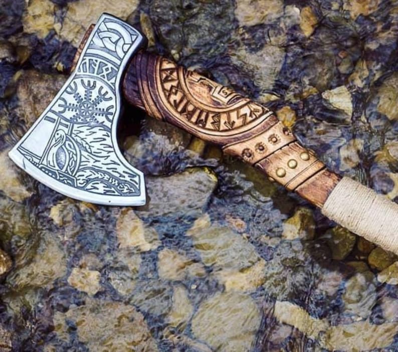 Puede incluir: Un hacha hecha a mano con mango de madera y cabeza de acero. La cabeza est&aacute; decorada con intrincados tallados, incluyendo un s&iacute;mbolo de una rueda solar y un p&aacute;jaro. El mango est&aacute; envuelto en cuerda y tiene una correa de cuero.