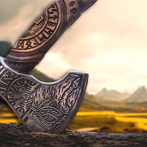 Puede incluir: Un hacha de plata con tallas intrincadas y un mango de madera con runas. El hacha est&aacute; apoyada sobre una superficie de madera con un fondo borroso de monta&ntilde;as y un campo.