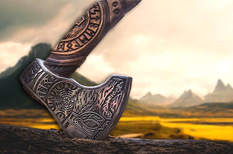 Puede incluir: Un hacha de plata con tallas intrincadas y un mango de madera con runas. El hacha est&aacute; apoyada sobre una superficie de madera con un fondo borroso de monta&ntilde;as y un campo.