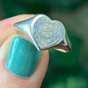 May include: A silver ring with a heart-shaped face. The heart has a textured design with a circular emblem in the center. The ring is held up by a person with turquoise nail polish.