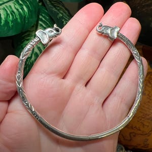 May include: A silver-toned open cuff bracelet featuring two sculpted elephant heads at the terminals. The band is decorated with an intricate, repeating etched pattern along its length, providing a textured appearance to the metallic surface.