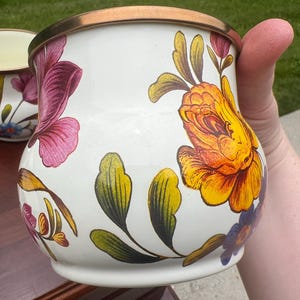 May include: A white enamel mug featuring a vibrant floral pattern with large yellow and orange roses, purple blossoms, and green leaves. The mug has a rounded, bulbous shape and a metallic bronze-colored rim along the top edge.