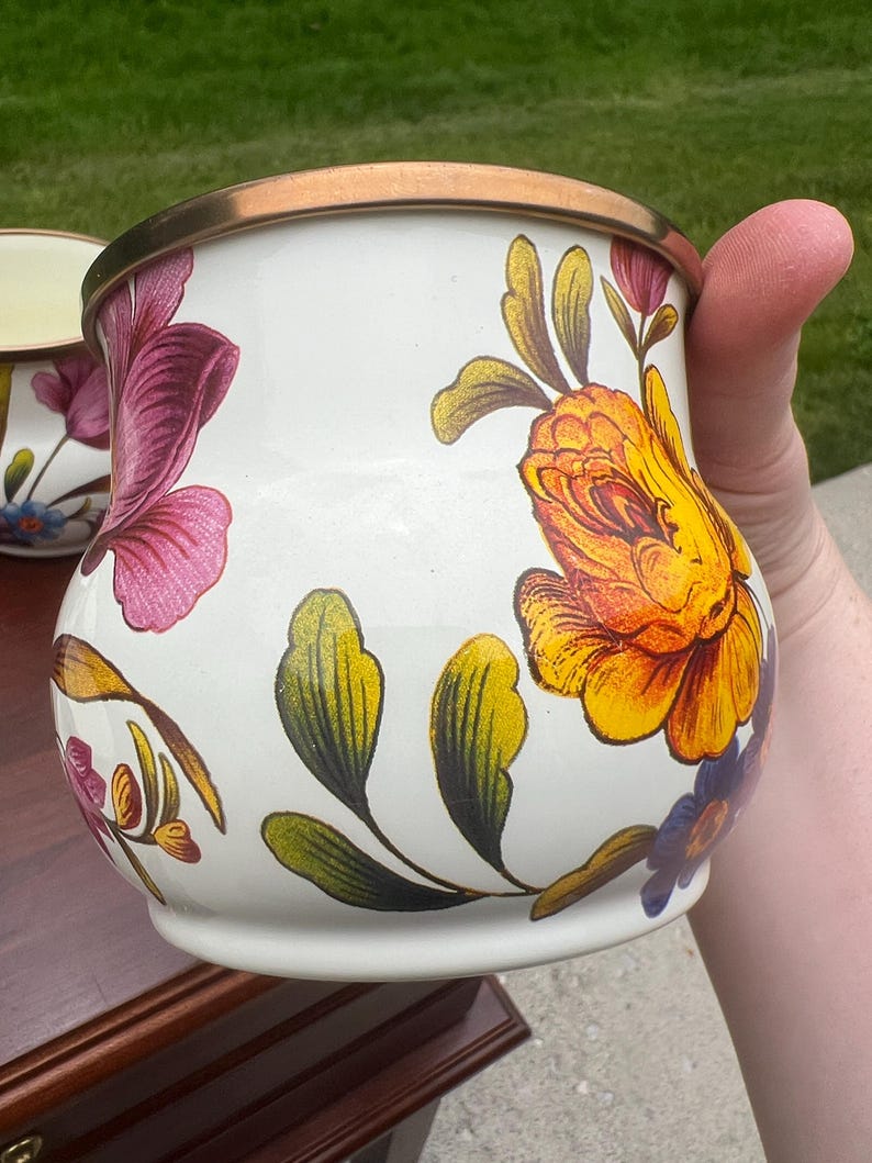 May include: A white enamel mug featuring a vibrant floral pattern with large yellow and orange roses, purple blossoms, and green leaves. The mug has a rounded, bulbous shape and a metallic bronze-colored rim along the top edge.