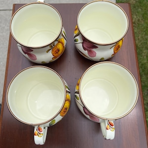 May include: A set of four cream-colored ceramic mugs with a floral pattern featuring yellow and purple flowers. Each mug has a rounded shape, a smooth interior, and a thin metallic-colored rim. The handles are decorated with small floral motifs.